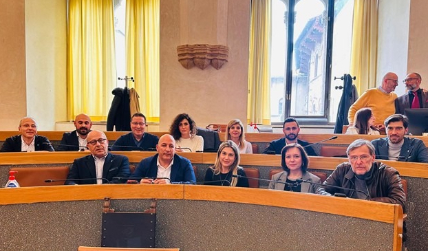 Group of individuals seated in a meeting room, some smiling and engaging, with large windows and yellow curtains in the background.