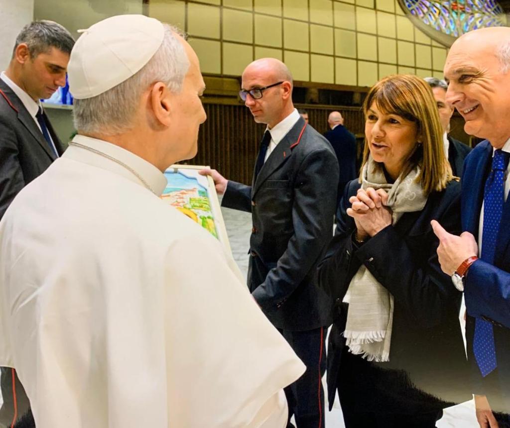 A group of people, including a religious leader, is engaged in conversation while a woman holds a piece of art in a formal setting.