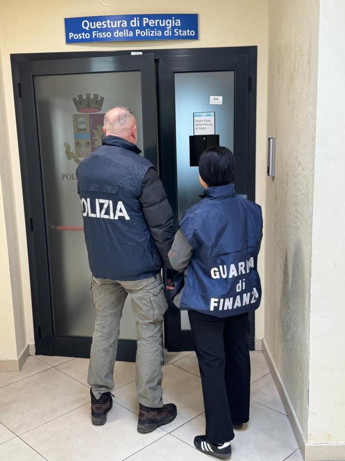 Two officers in jackets labeled 'POLIZIA' and 'GUARDIA di FINANZA' standing in front of a door at the Questura di Perugia police station.