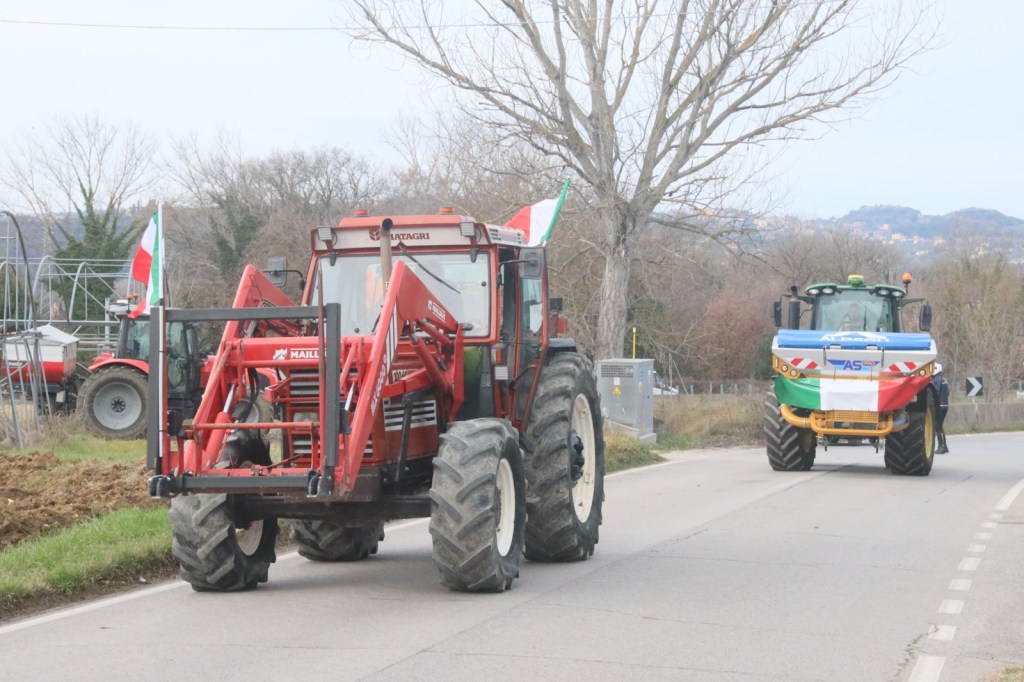 A red tractor with a front loader and Italian flags drives along a rural road, followed by a colorful tractor with a flag design. Trees and agricultural equipment can be seen in the background.
