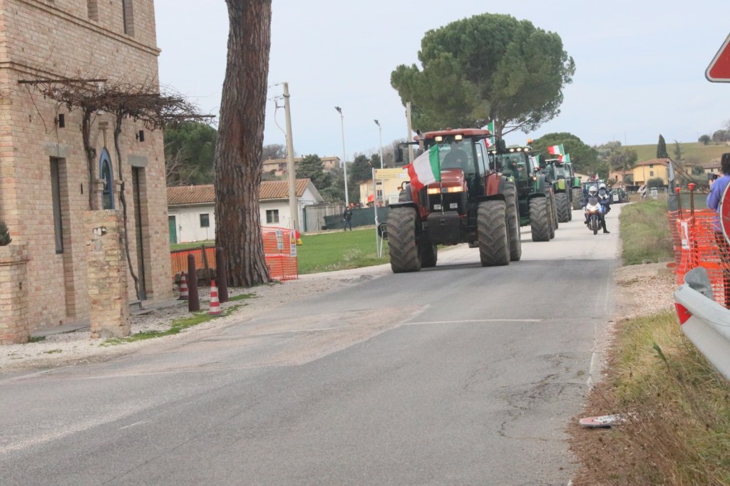 A line of tractors decorated with Italian flags driving down a rural road, with a brick building and trees in the background.