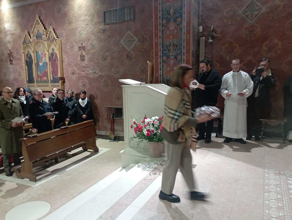 A group of people in a church setting, with a woman walking in front while holding a package. Clergy members stand at the altar, and attendees are gathered in formal attire, some holding booklets.