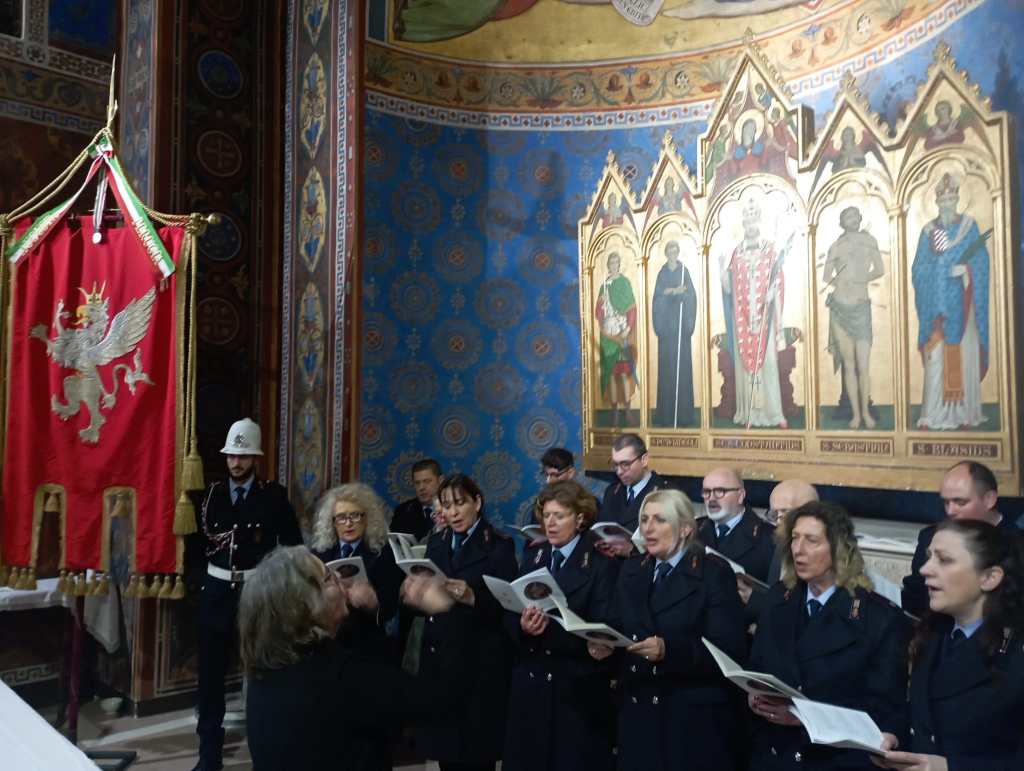 A choir of performers in formal attire singing in front of religious artwork inside a decorated hall, with a red flag featuring a golden eagle in the foreground.