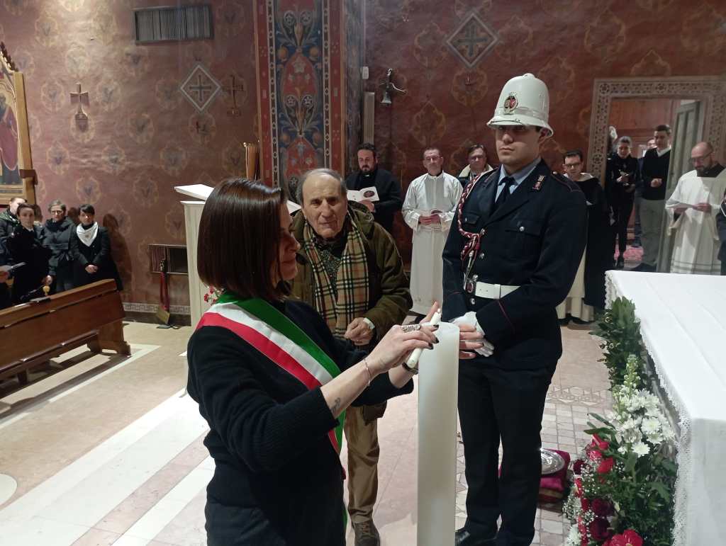 A woman in official attire, holding a scroll, stands in a church surrounded by a diverse group of attendees, including a police officer in uniform. The interior features decorative walls and attendees in the background observing the ceremony.