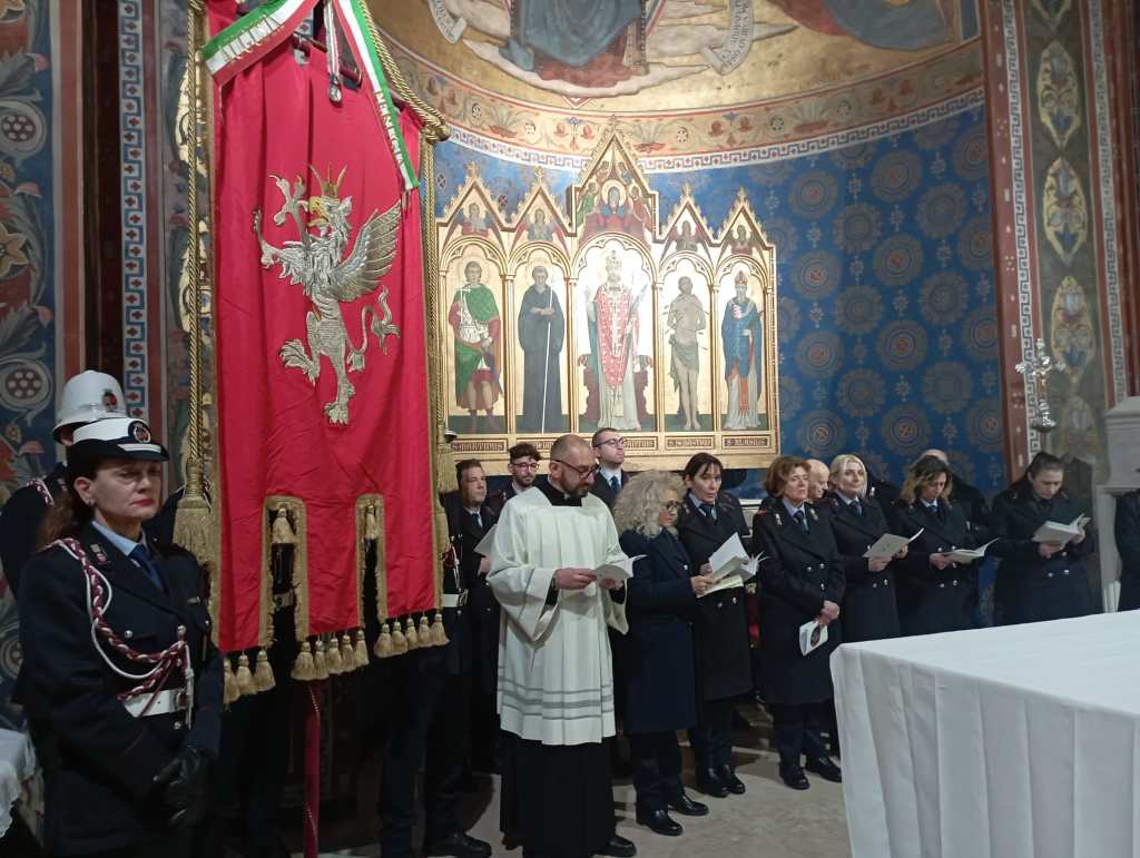 A ceremonial gathering in a decorated chapel featuring people in uniform and a large red banner with a griffin. They are participating in a religious service, led by a figure in white robes, with a backdrop of religious artwork.