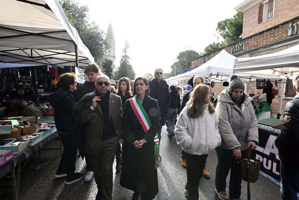 A group of people walking through a market, with stalls selling various items. One woman in the foreground, wearing a sash, appears to be a public official, while others browse nearby.