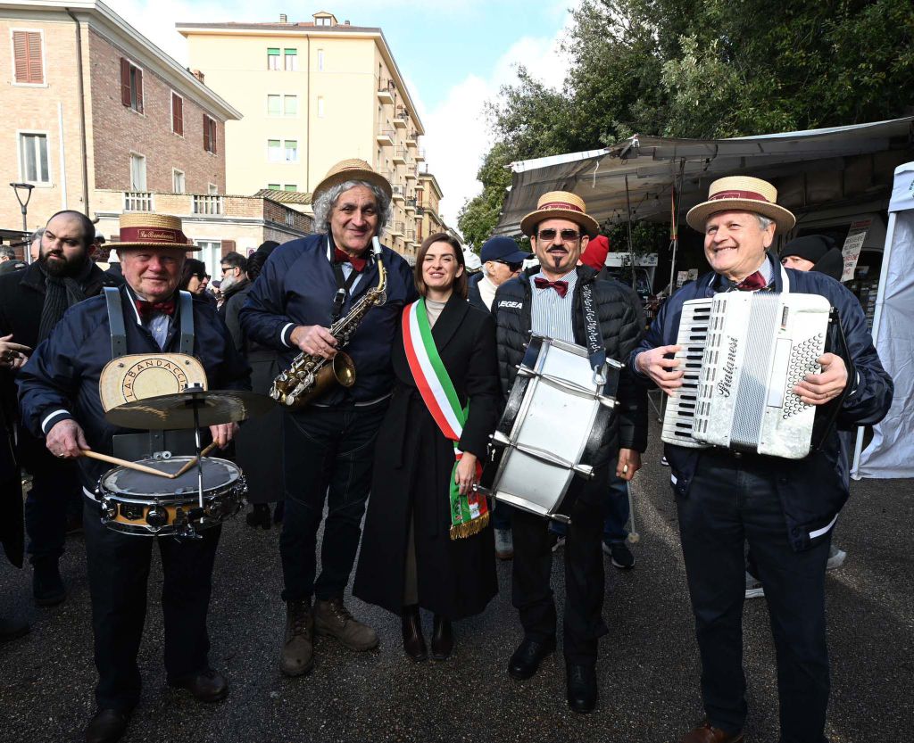 A group of five musicians in traditional attire, including hats and bow ties, posing for a photo outdoors. They play various instruments: a saxophone, drum, and accordion. A woman in the center, wearing a black coat and a sash with green, white, and red stripes, stands among them. The background features buildings and a market setting.