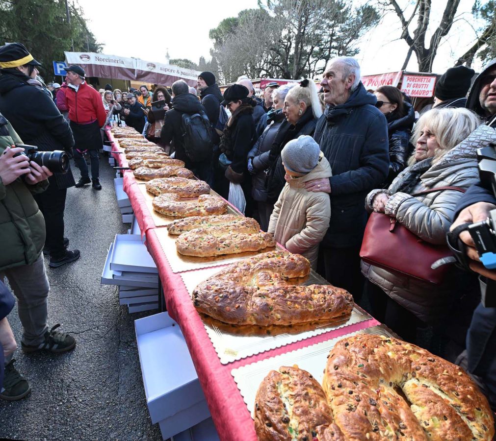 A group of people gathered at a market, admiring a long table filled with large, decorative breads. Some individuals are taking photos, while others are chatting excitedly.