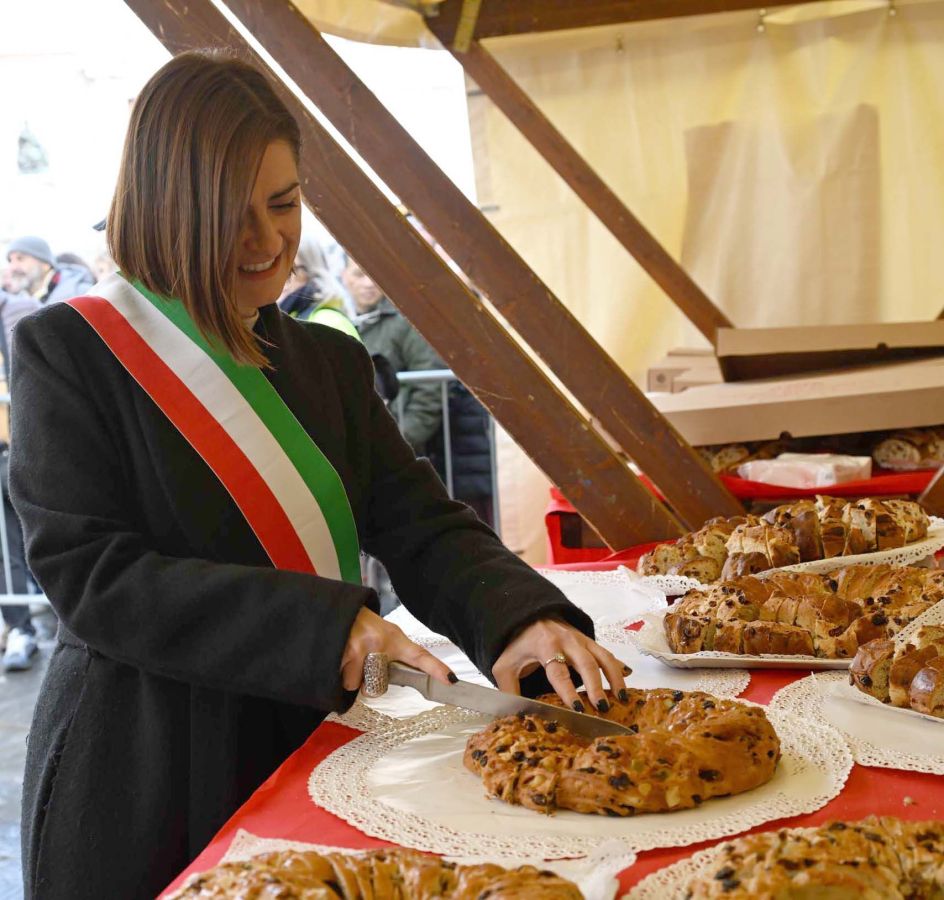 A woman wearing a ceremonial sash slices a round pastry on a table decorated with lace doilies, surrounded by various baked goods in a festive setting.