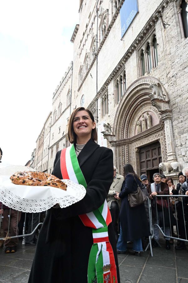 A woman wearing a sash holds a large traditional cake in front of a historic building, with a crowd of people in the background.