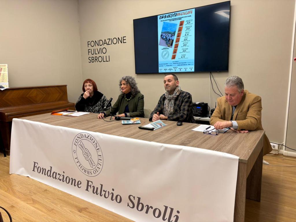 A panel discussion at Fondazione Fulvio Sbrolli, featuring four individuals seated at a table with notes, a banner in front, and a projector screen displaying event information.