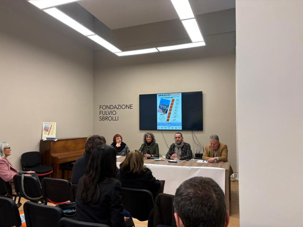 A panel discussion taking place at Fondazione Fulvio Sbroli, featuring four speakers seated at a table with a presentation displayed on a screen in the background.