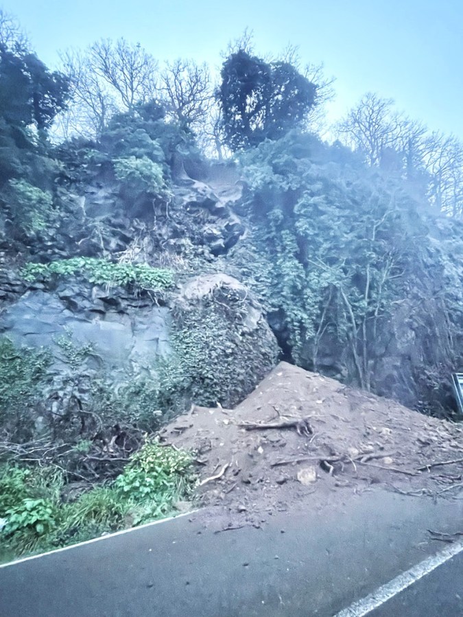 A landslide covering part of a road with rocky terrain and overgrown vegetation in a misty environment.