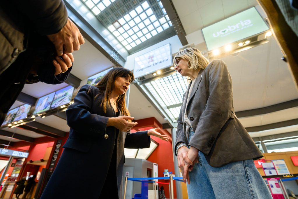 Two women engaged in conversation at a travel or airport setting, with screens displaying information in the background.