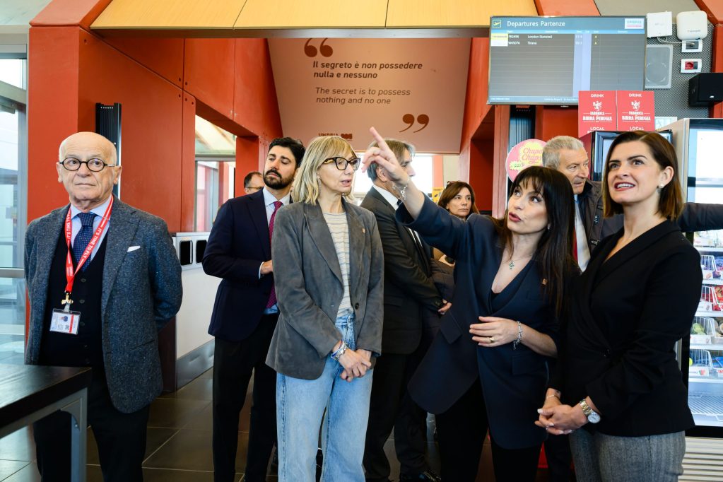A group of people engaged in conversation inside a modern building, with one woman pointing towards a display screen while others listen attentively.