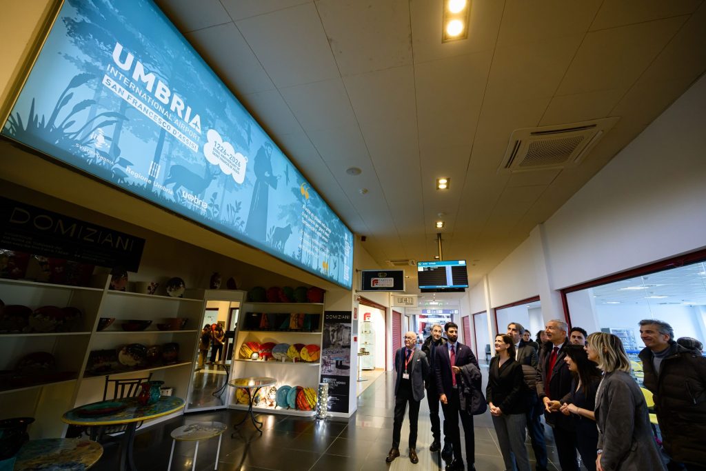 Interior view of Umbria International Airport featuring a digital display and a group of people in business attire, with colorful ceramic items on display.