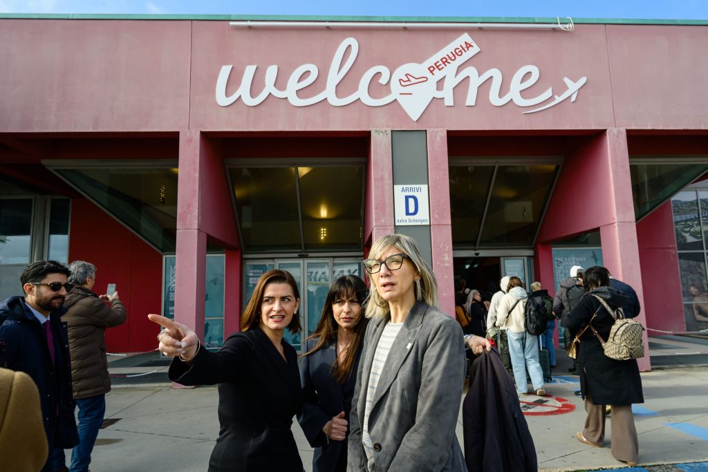 Three women standing outside an airport terminal with a sign that says 'welcome Perugia' in the background.