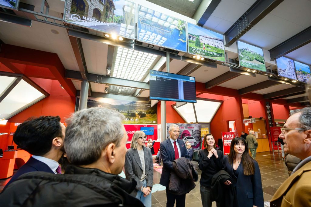 A group of people interacting in a busy airport terminal decorated with digital display screens showcasing different tourist destinations.