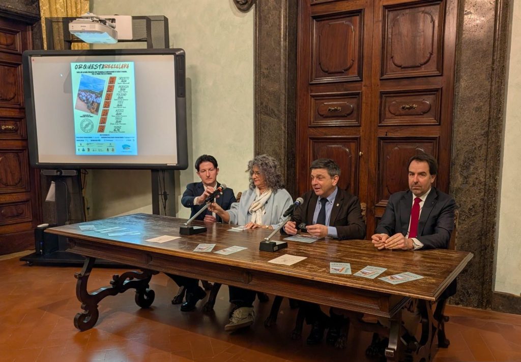 A press conference featuring four speakers seated at a table, with a projector display in the background showing event information. The setting includes wooden decor and a formal atmosphere.