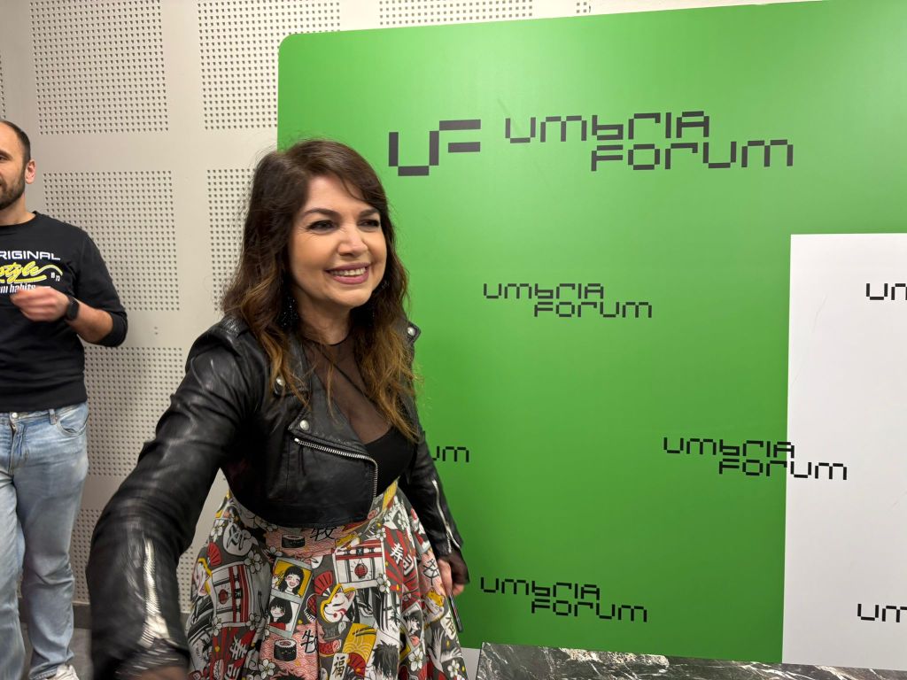 A smiling woman with brown hair wearing a black leather jacket and a colorful skirt stands in front of a green backdrop with 'Umbria Forum' written on it. A man in a black shirt is visible in the background.