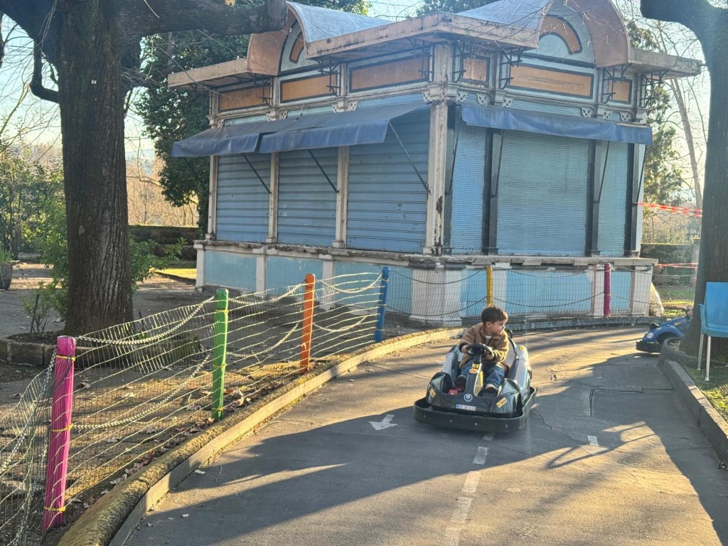 A child driving a go-kart on a track, with a closed amusement park booth in the background and colorful fence posts along the path.