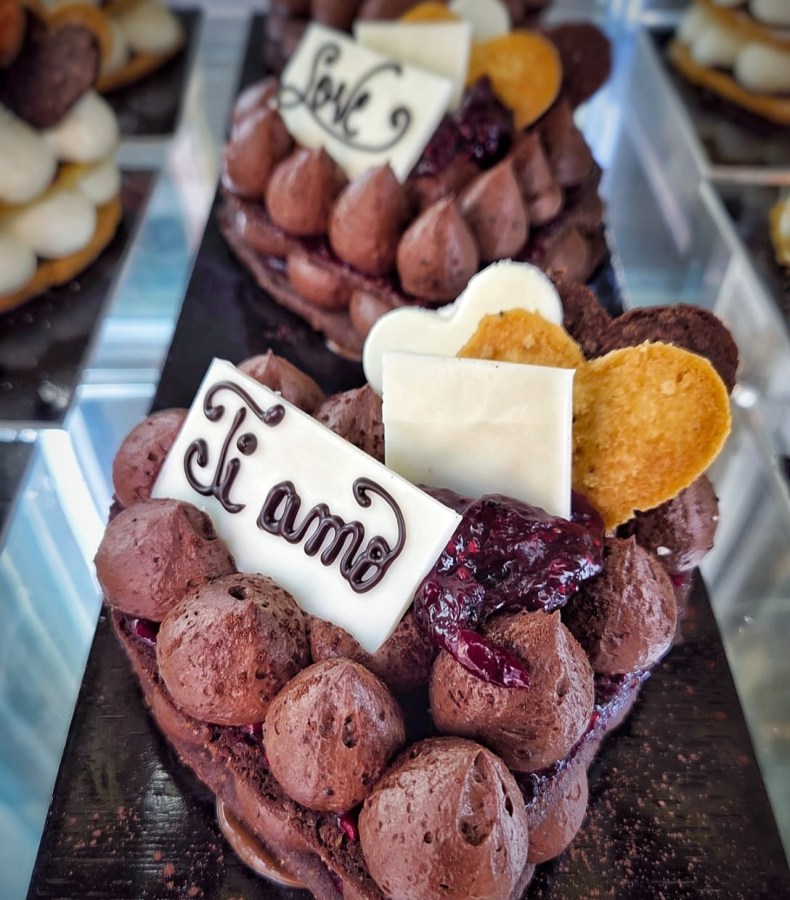 A heart-shaped chocolate cake decorated with chocolate mousse, a white chocolate square, and cookies, featuring the phrases 'Ti amo' and 'Love'.