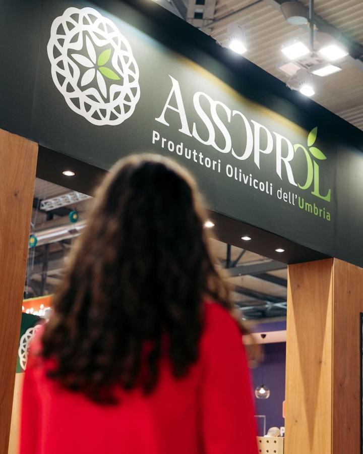 A woman with curly hair stands in front of a booth displaying the ASSOPROL logo, which represents olive oil producers from Umbria, Italy.