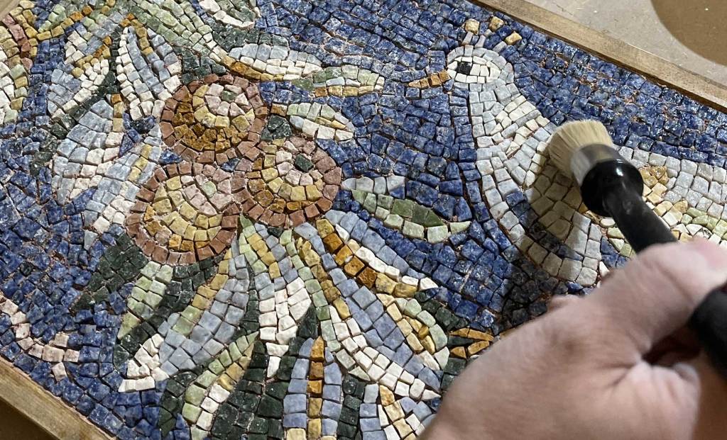 Close-up of a hand brushing a colorful mosaic art piece featuring flowers and a bird.