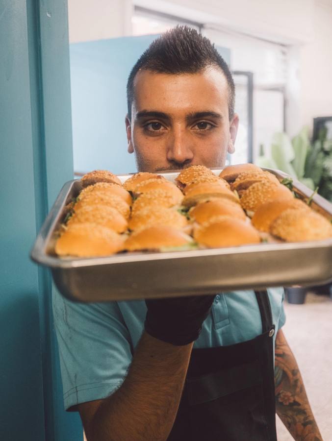 A chef holding a tray of hamburger sliders with sesame seed buns, smiling and standing against a blue wall.
