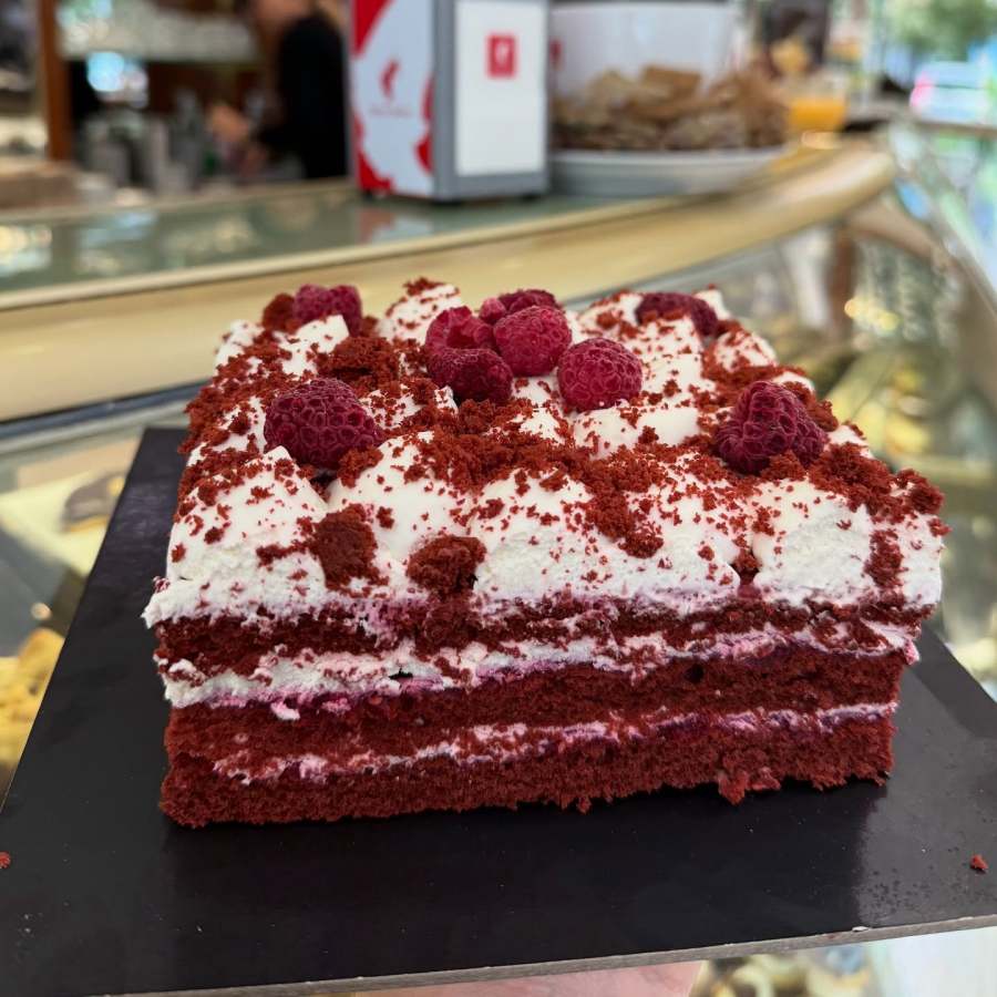 A slice of red velvet cake topped with whipped cream and fresh raspberries, displayed on a black plate.