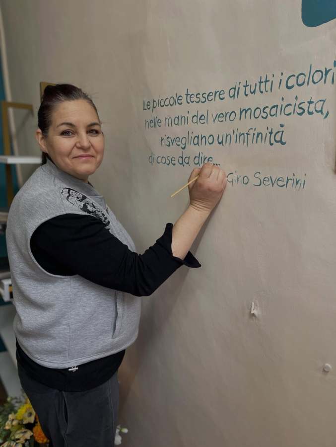 A woman wearing a sleeveless gray top is painting a quote on a beige wall, holding a brush in her right hand and smiling at the camera.