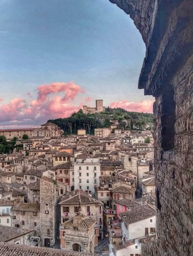 A scenic view of a historic town with stone buildings and tiled roofs, framed by an archway. In the background, a castle sits atop a hill under a colorful sky with pink clouds.