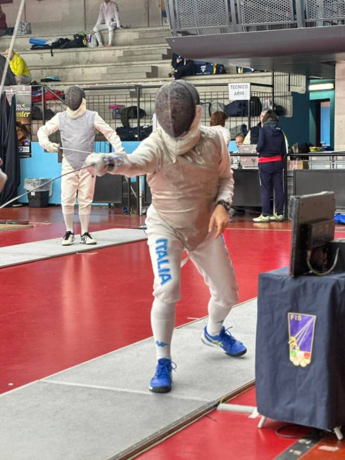 A fencer in full gear, wearing a mask and protective clothing, lunges with a sword on a red indoor fencing strip. The opponent is positioned in the background, while spectators and officials are visible in the stands.
