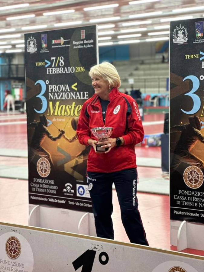 A woman wearing a red jacket holds a trophy while standing in front of a promotional banner for a national masters fencing competition. The background features a sports hall with fencing mats.