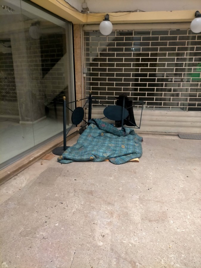 A makeshift bed with a blue plaid blanket is set up on a concrete floor beside a window displaying a brick wall in the background. Two chairs are nearby, one of which is overturned.