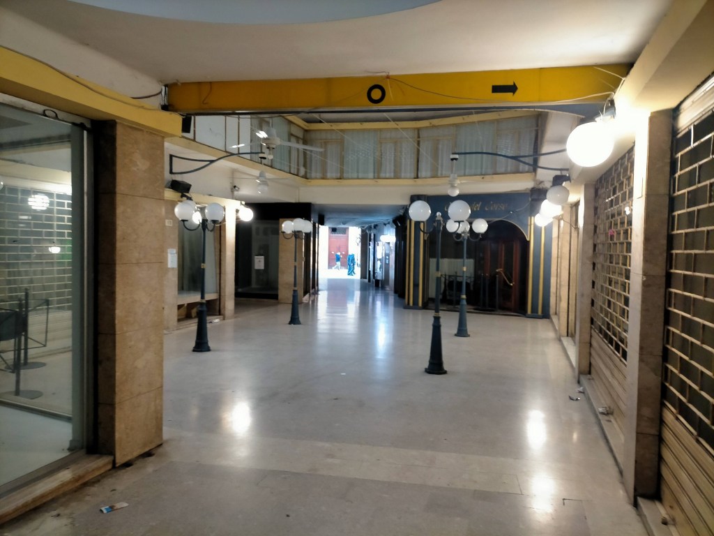 A wide view of an empty corridor lit by street lamps, with storefronts on the sides and a staircase in the background.