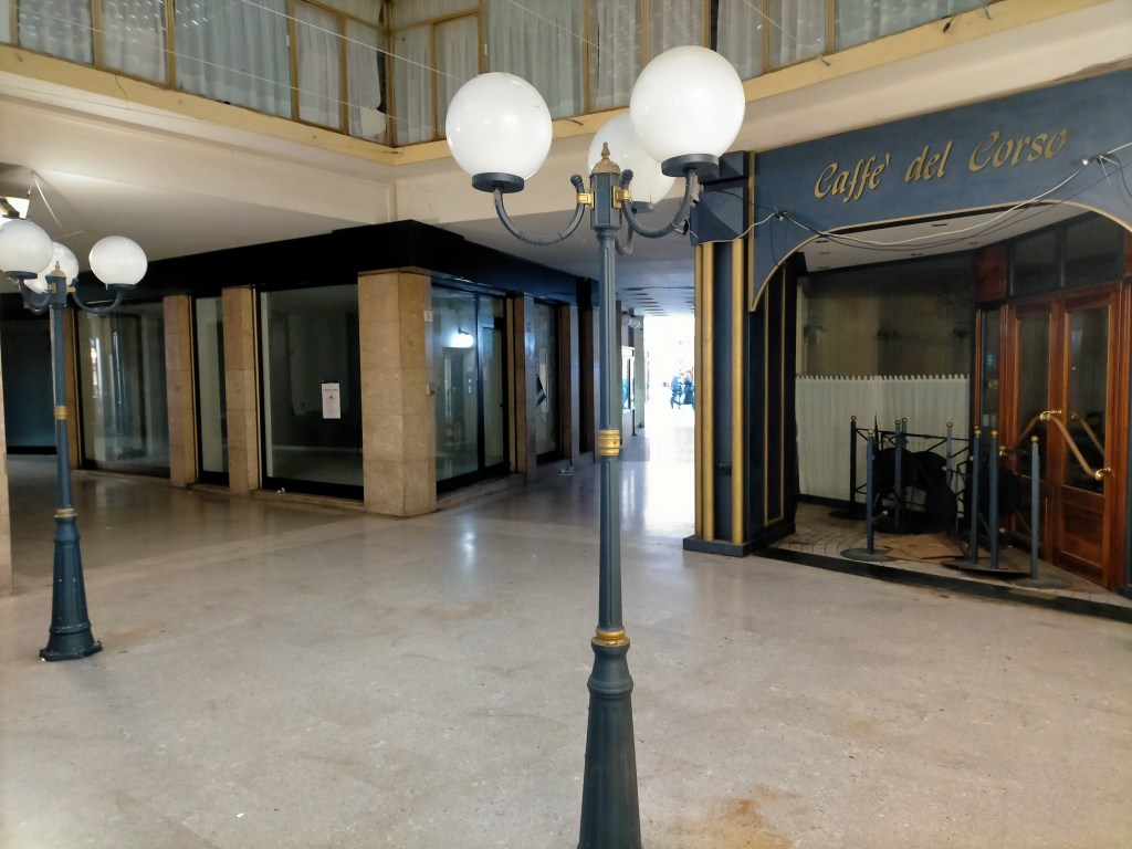 Interior of a shopping arcade featuring decorative street lamps and a cafe entrance, with empty storefronts in the background.