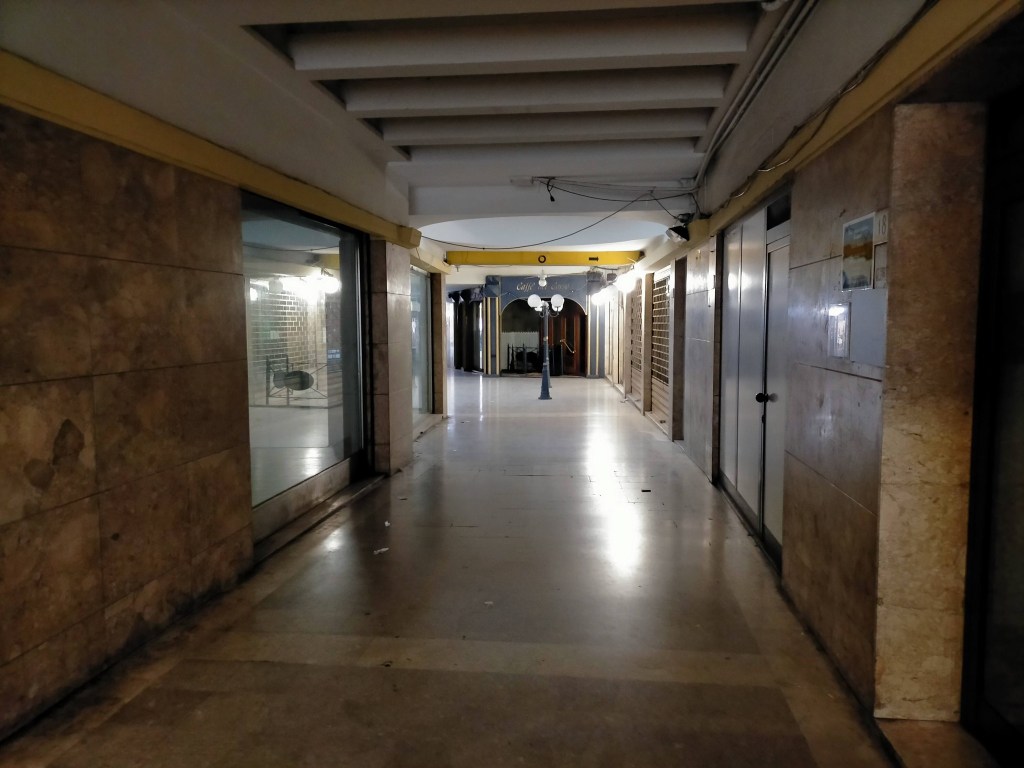 A dimly lit corridor with stone walls, featuring closed shopfronts and a decorative lamp post. The floor is smooth and shiny, leading to an open area in the distance.