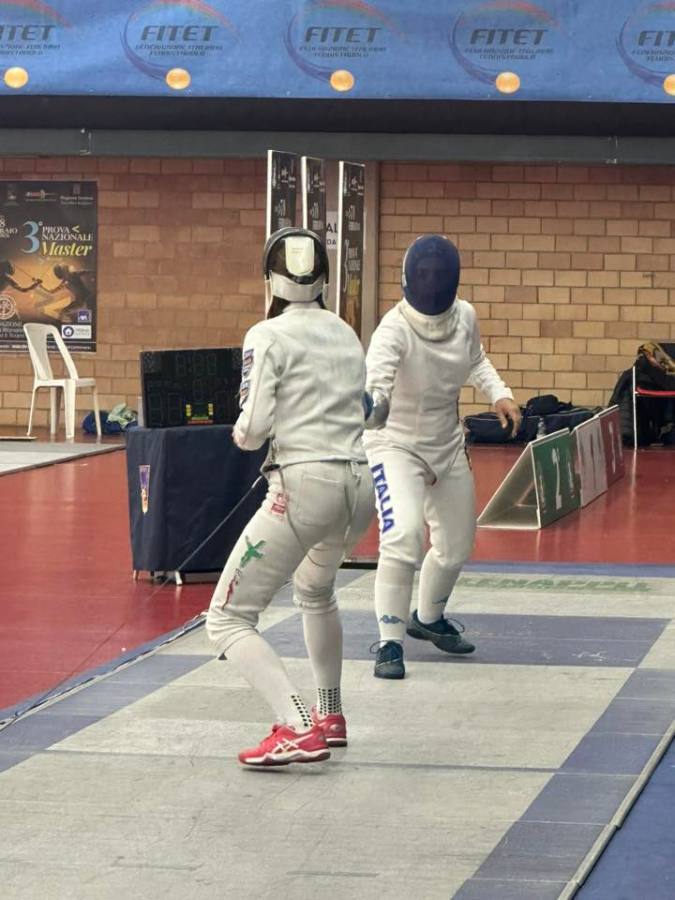 Two fencers engaged in a duel on a fencing piste, wearing protective gear and masks, with a scoreboard and banners in the background.