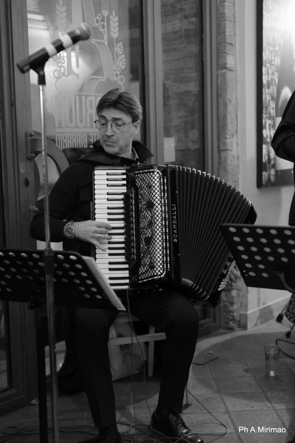 A musician playing an accordion on stage, wearing glasses and a black outfit, with a microphone and music stand in the foreground.