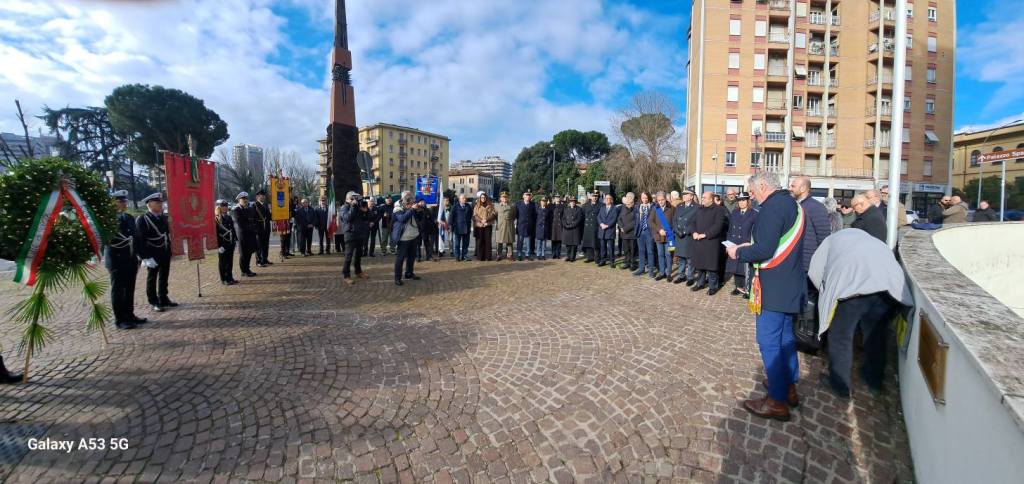 A gathering of people at a commemorative event in a public square, with officials in formal attire, including one wearing a tricolor sash, addressing the crowd. The scene includes a decorative wreath and various flags in the background, along with a mix of trees and buildings under a partly cloudy sky.