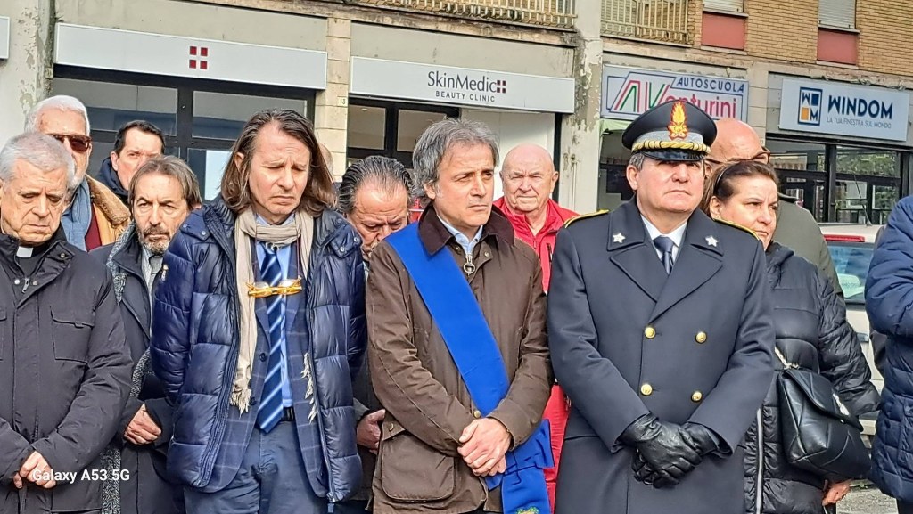 A group of people, including officials in formal attire, standing solemnly during a ceremony or memorial event. Some individuals are wearing coats, and the atmosphere appears serious and respectful.