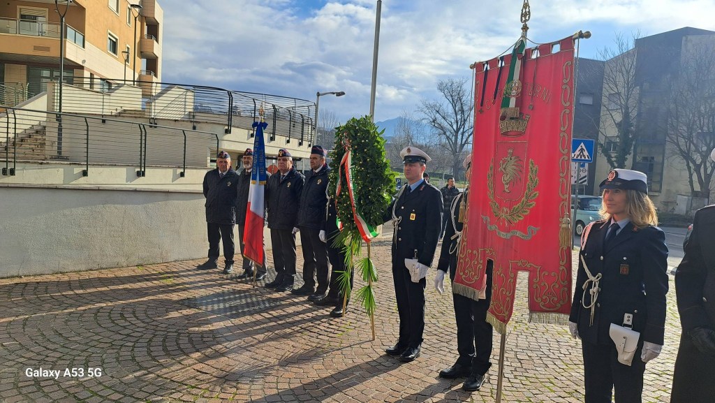 A group of individuals in uniform during a commemorative event, standing in front of a large flag and a ceremonial wreath. The Italian flag is displayed, and the scene is set against a backdrop of buildings and a partly cloudy sky.