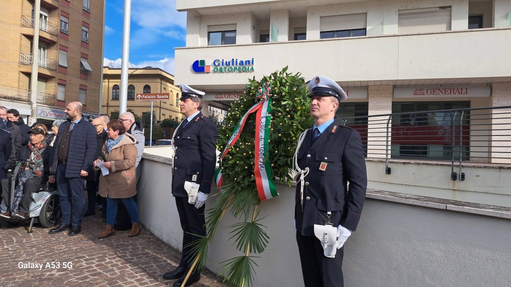 A group of people gathered outside a building, some holding papers, while two officers in formal attire stand by a wreath decorated with the Italian flag. An elderly man in a wheelchair is visible in the crowd.