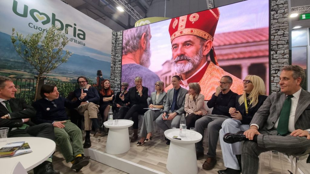 A panel discussion featuring several people seated around a white table, with a large backdrop displaying the logo 'Umbria' and an image of a man wearing a ceremonial headdress.