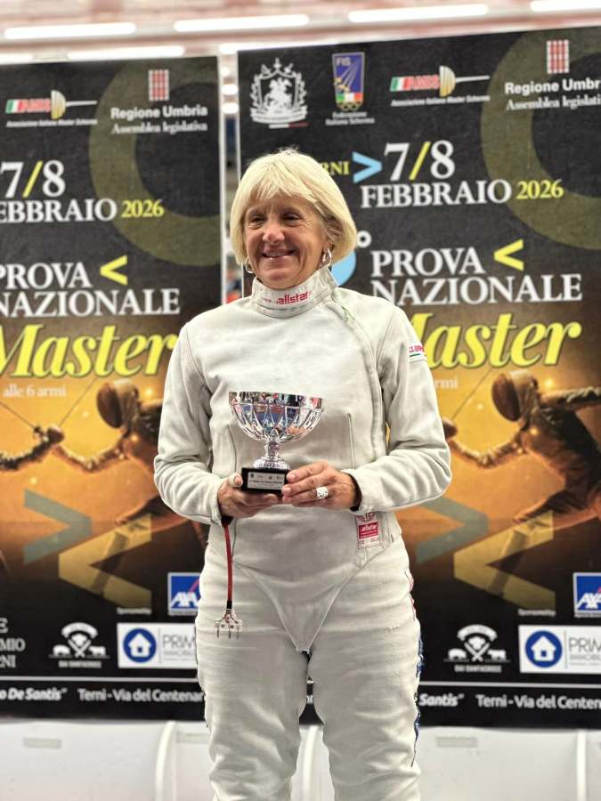 A smiling female fencer in a white fencing uniform holds a trophy, standing in front of promotional banners for a fencing competition.