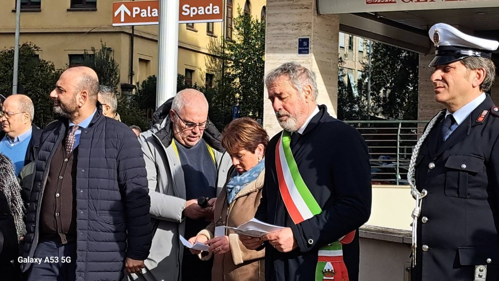 A group of people gathered outdoors, with a man wearing a ceremonial sash speaking and several individuals, including women and men, listening attentively. Behind them, a sign points to 'Pala Spada'.