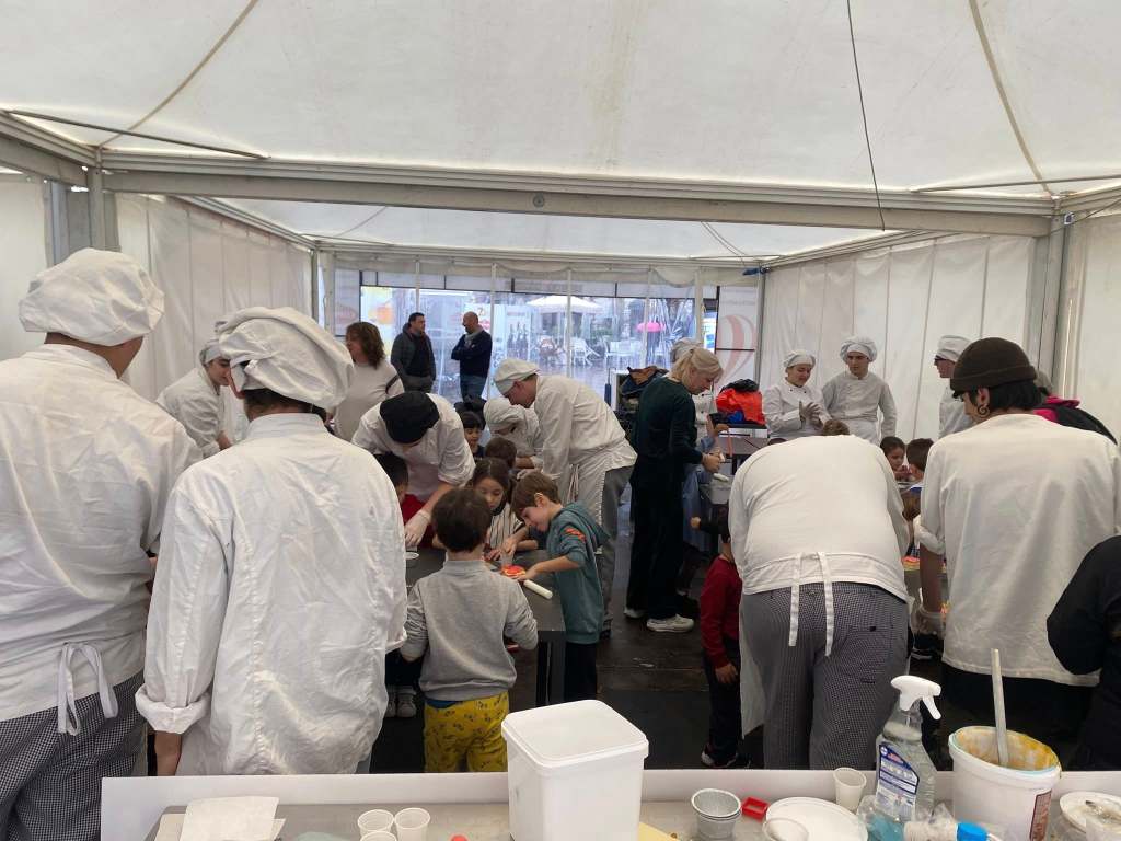 A busy cooking class taking place inside a tent, with chefs in white uniforms and children engaged in food preparation activities.
