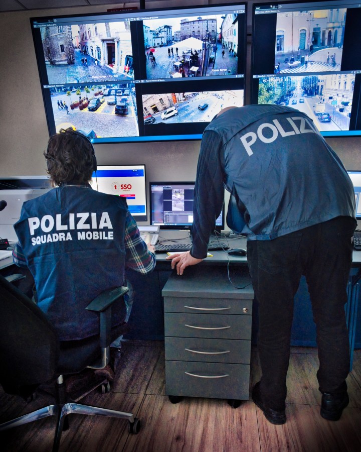 Two police officers monitoring multiple surveillance screens in a control room, displaying various outdoor locations.