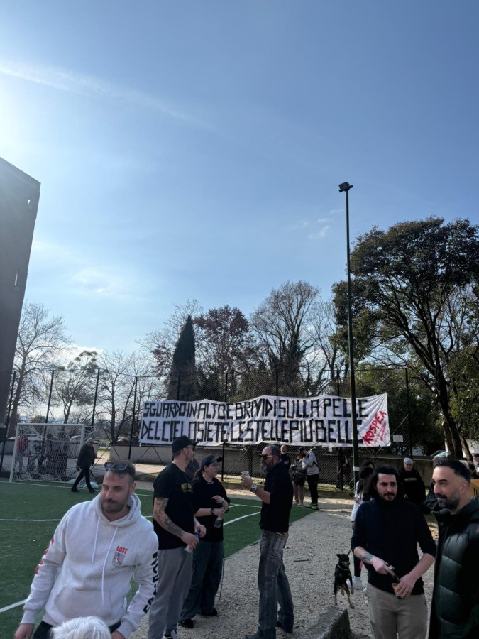 A group of people gathered in a park with a large banner that reads a message in Italian, set against a clear blue sky. There are trees and a sports field in the background.